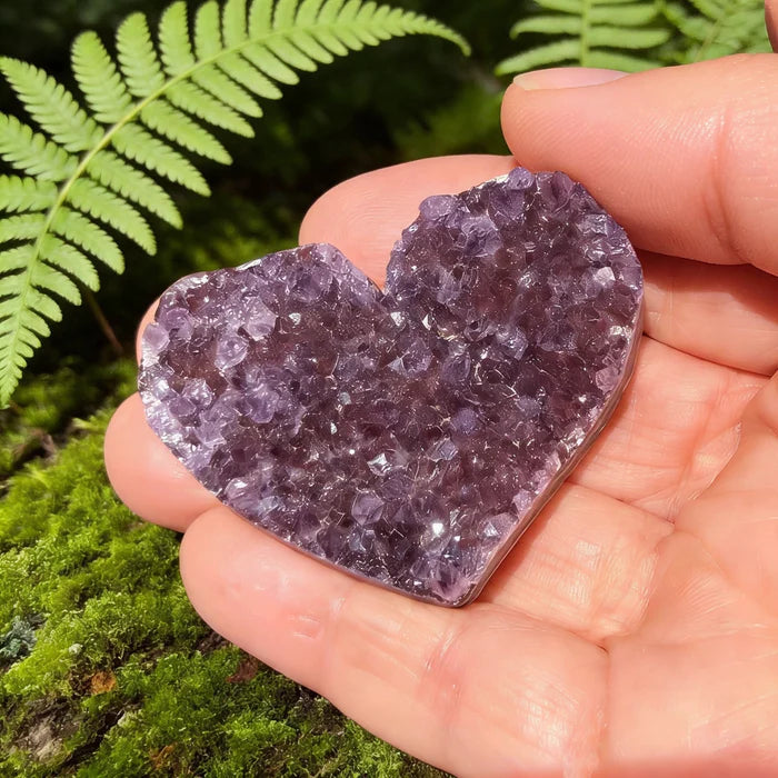 Sparkling purple amethyst cluster heart resting in an open palm beside a fern