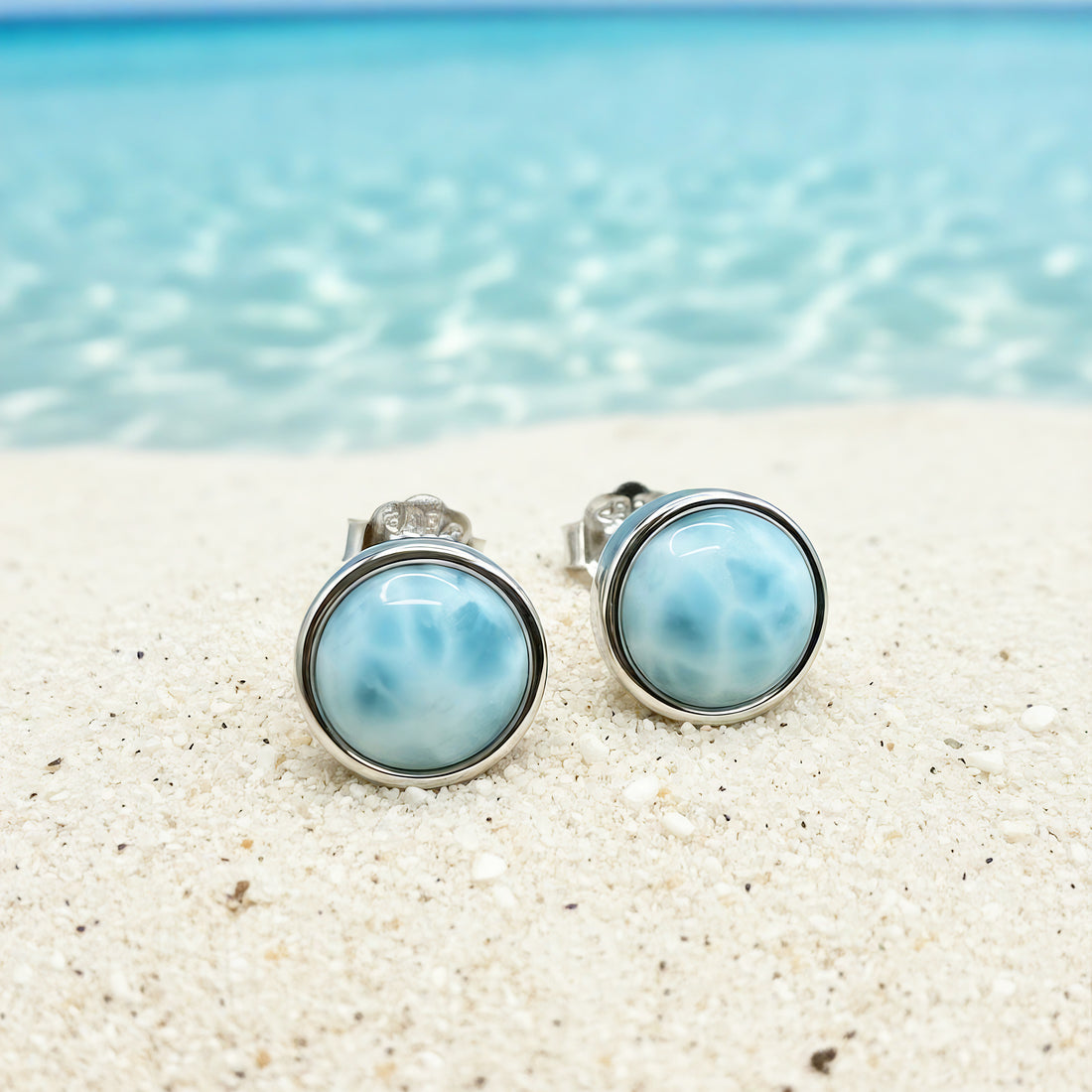 A pair of round Larimar stud earrings in polished silver bezels resting on white sand by the sea