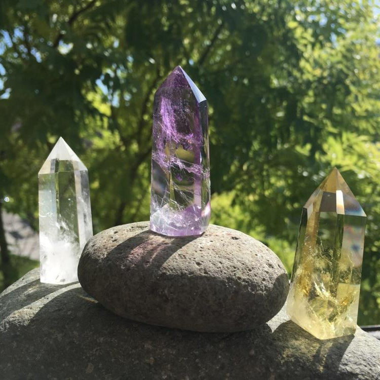 A clear quartz point, an amethyst point and a citrine point standing on smooth gray stones in sunlight