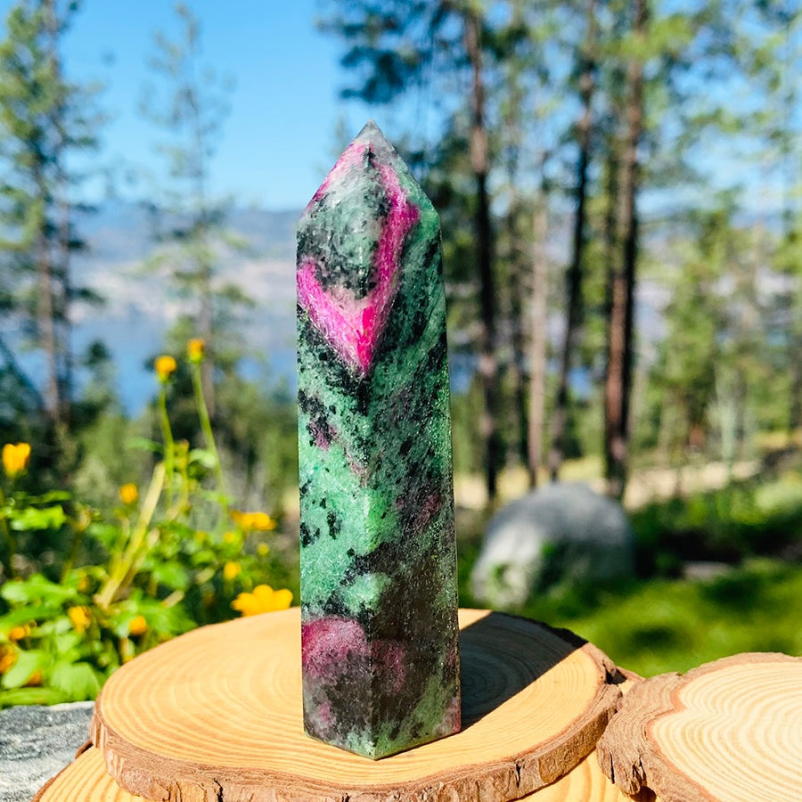A ruby in zoisite point, green with black webbing and bright pink ruby patches, standing on a wooden round among pines and yellow flowers