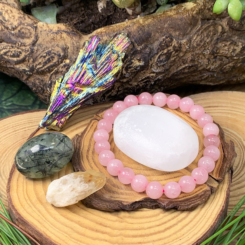 The rose quartz mala bracelet around a white selenite ellipse on a wood slice, with an iridescent peacock kyanite fan, a green prehnite ellipse and a pale citrine shard