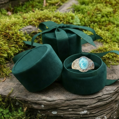 The larimar ring nestled in an open round green velvet jewelry box tied with ribbon, set on mossy stone