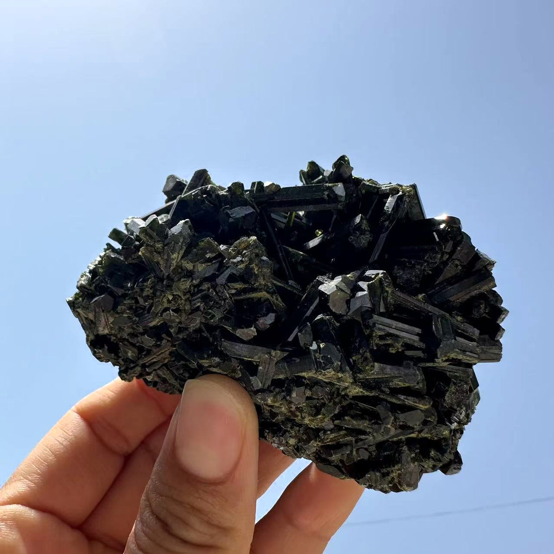 The green tourmaline specimen held up against a blue sky, a crisscross of glossy dark crystal blades flecked with green