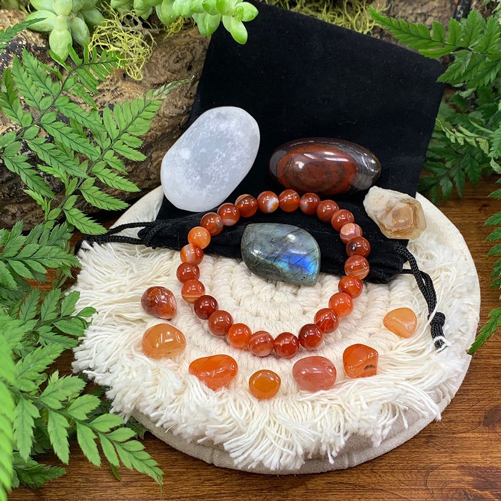 The striped red agate mala bracelet around a blue-flashing labradorite cabochon on a black velvet pouch, with a white selenite ellipse, a red tiger's eye ellipse, a pale citrine shard and seven orange carnelian mini gemstones