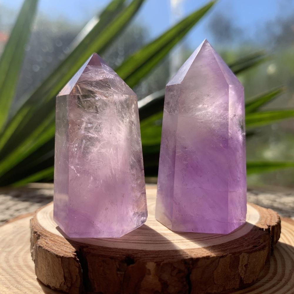 Two short, thick polished amethyst generators in lavender and violet standing on a wood slice in sunlight