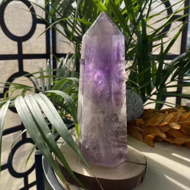 An amethyst tower standing on a wood slice beside palm fronds, its purple zones glowing through clear quartz