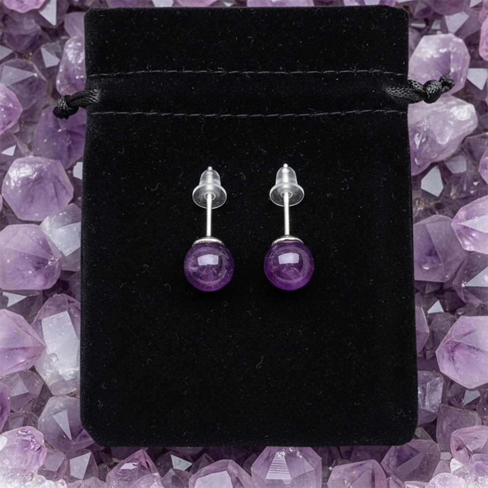 The pair of amethyst studs with their posts and clear backs, laid on a black velvet drawstring pouch among amethyst points