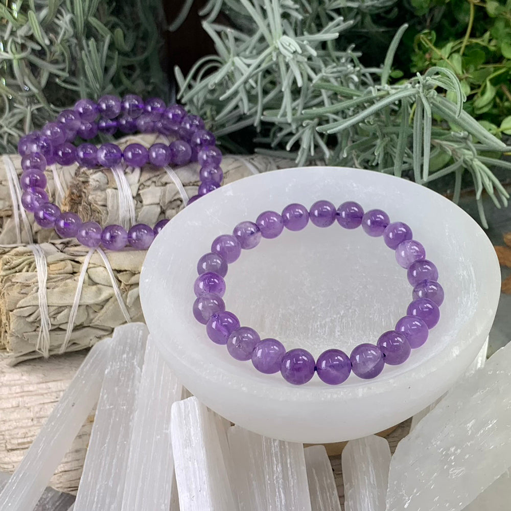 An amethyst bead bracelet resting in a white selenite bowl, with more bracelets on sage bundles behind