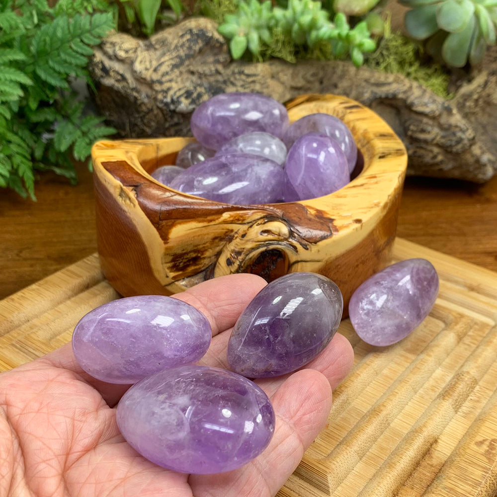 Three amethyst tumbled stones resting in an open palm in front of a wooden bowl of more amethyst