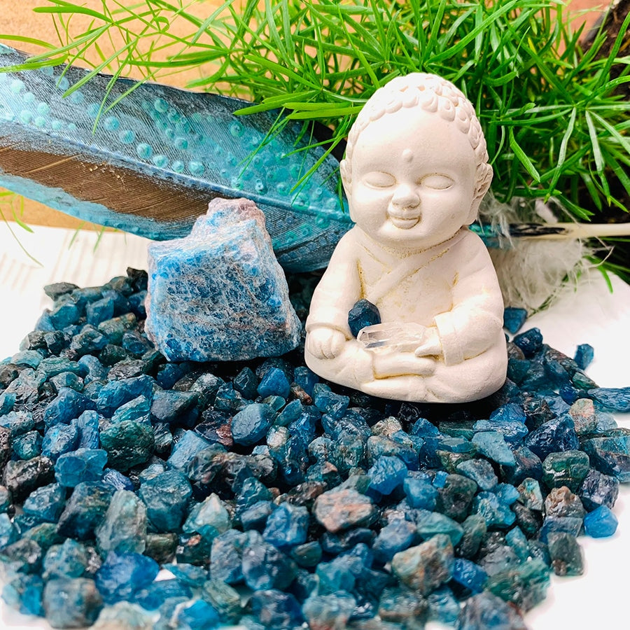 A bed of rough apatite mini gemstones around a larger apatite piece and a small Buddha figurine