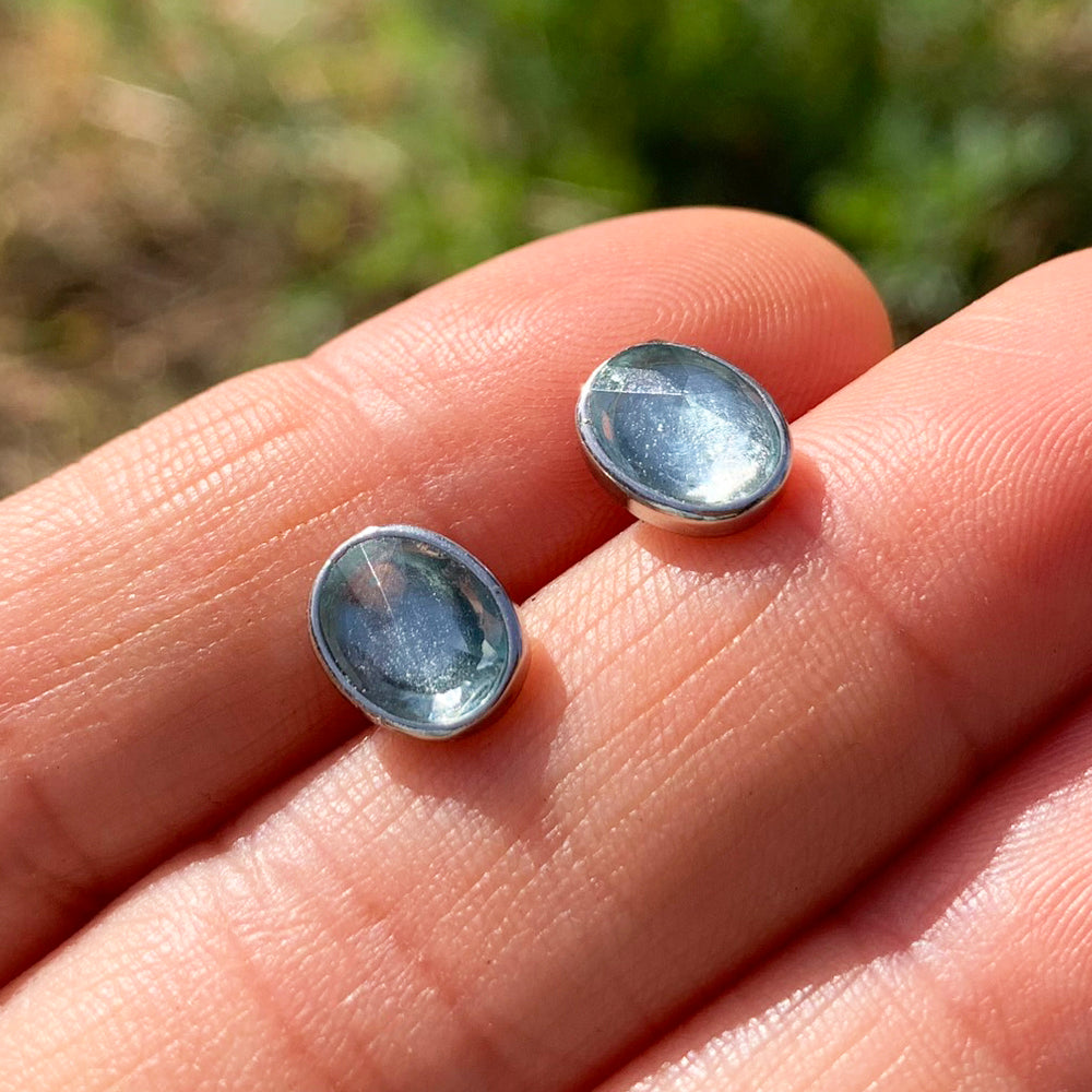 Two oval faceted aquamarine studs in sterling silver bezels resting on fingertips in sunlight