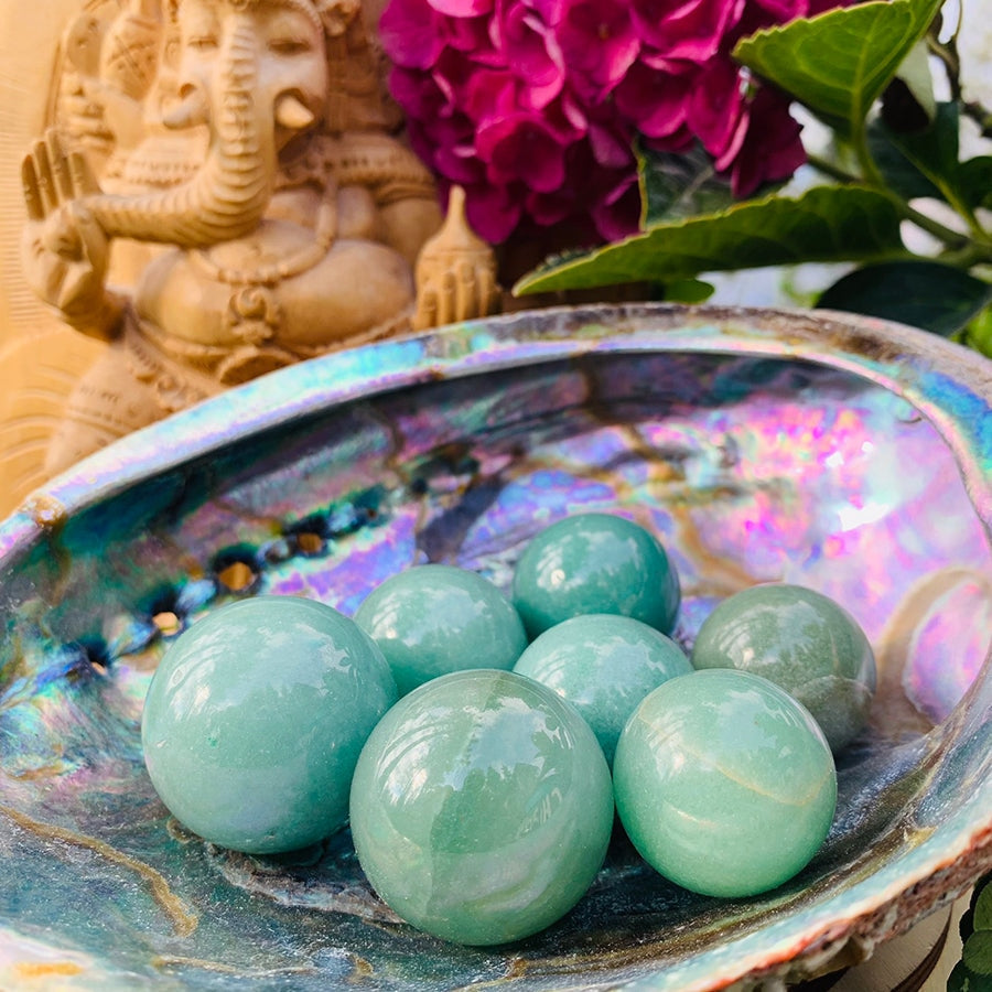 Several small green aventurine spheres in an iridescent abalone shell, with a carved statue and pink flowers behind as styling