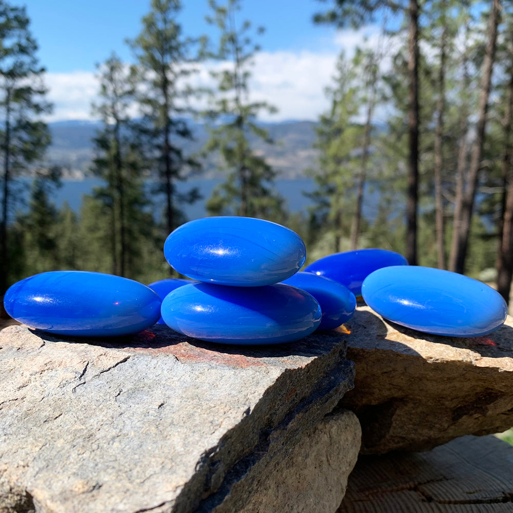 Several smooth blue Tranquil Azure palmstones stacked on sunlit rocks above a lake and pine trees