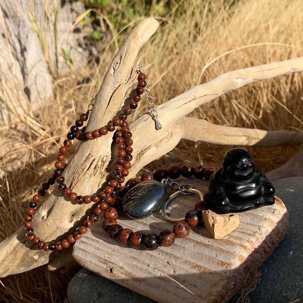 A red obsidian bead necklace and mala bracelet, a hematite keychain and a black agate Buddha arranged on driftwood in the sun