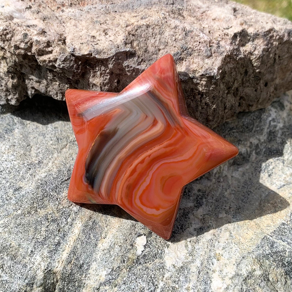 A polished five-pointed carnelian star in orange and red with cream and gray banding, resting on a rough rock in sunlight