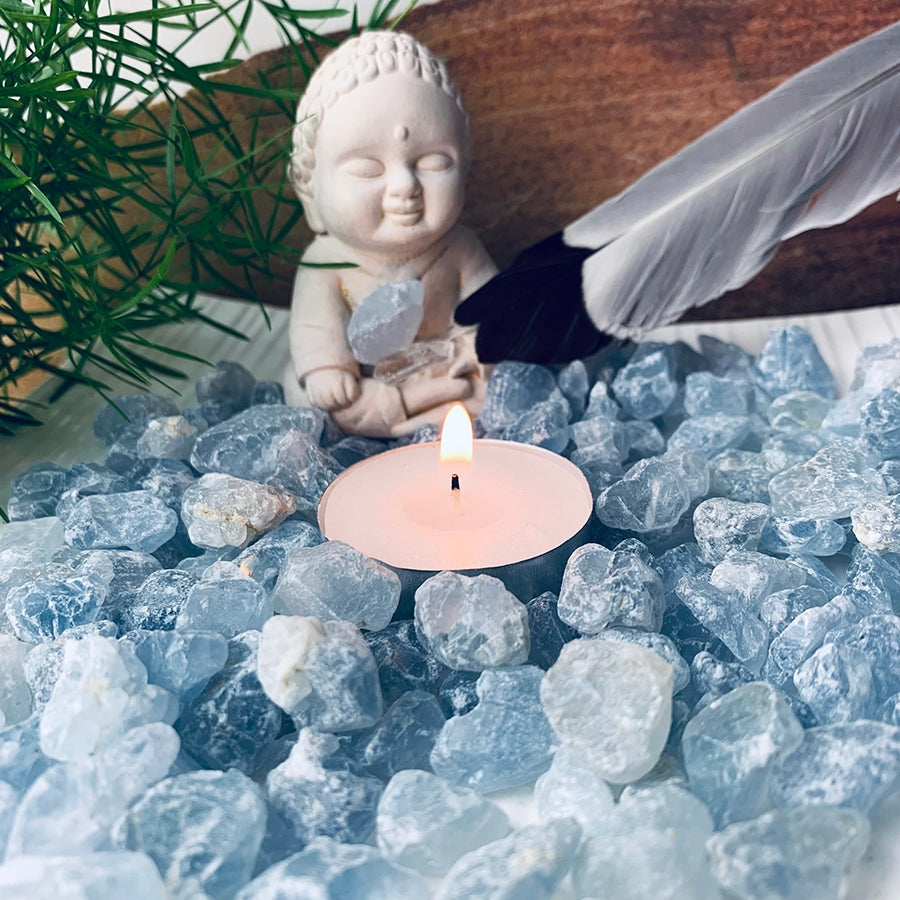Rough celestite stones spread around a lit tealight and a small seated figurine, styled with a feather