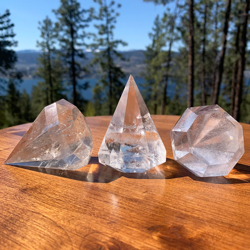Three faceted clear quartz diamond-cut crystals catching sunlight on a wooden table, with a forest and lake behind