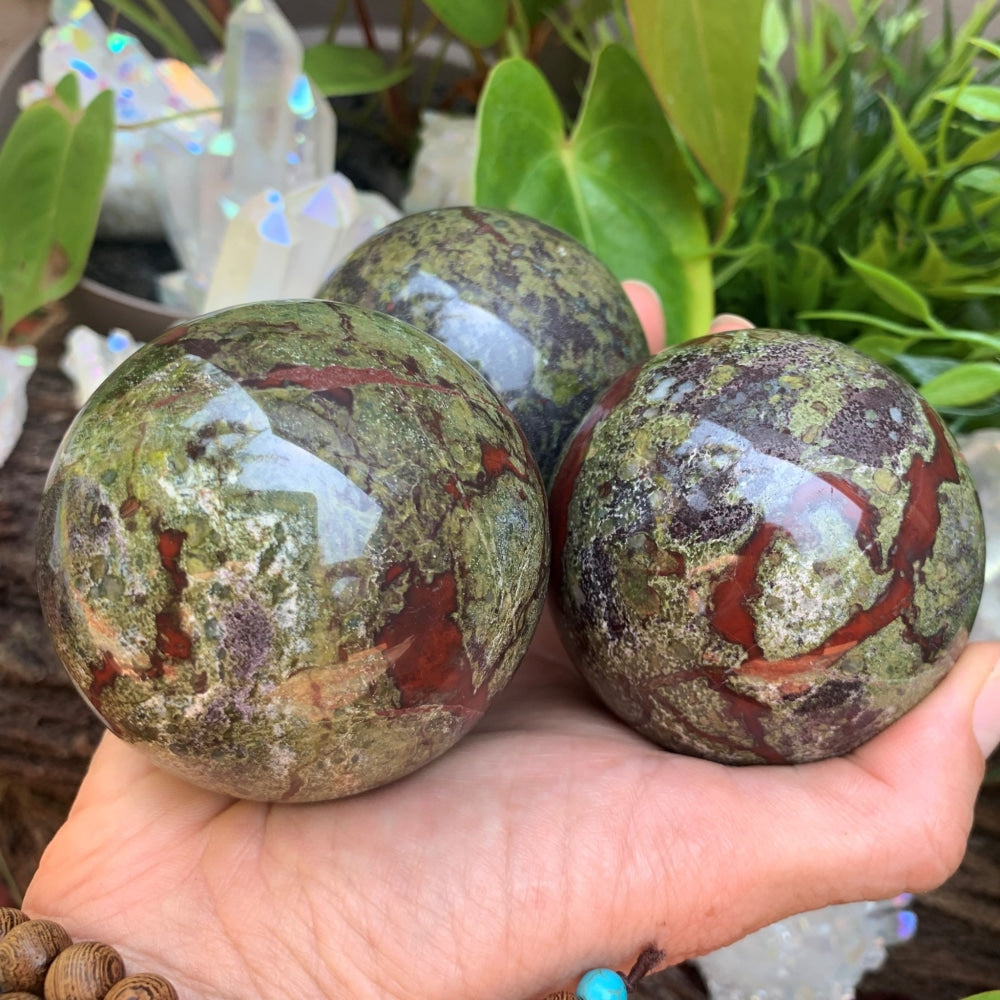 Three polished dragon blood jasper spheres with deep red veins through mossy green, held together in an open palm