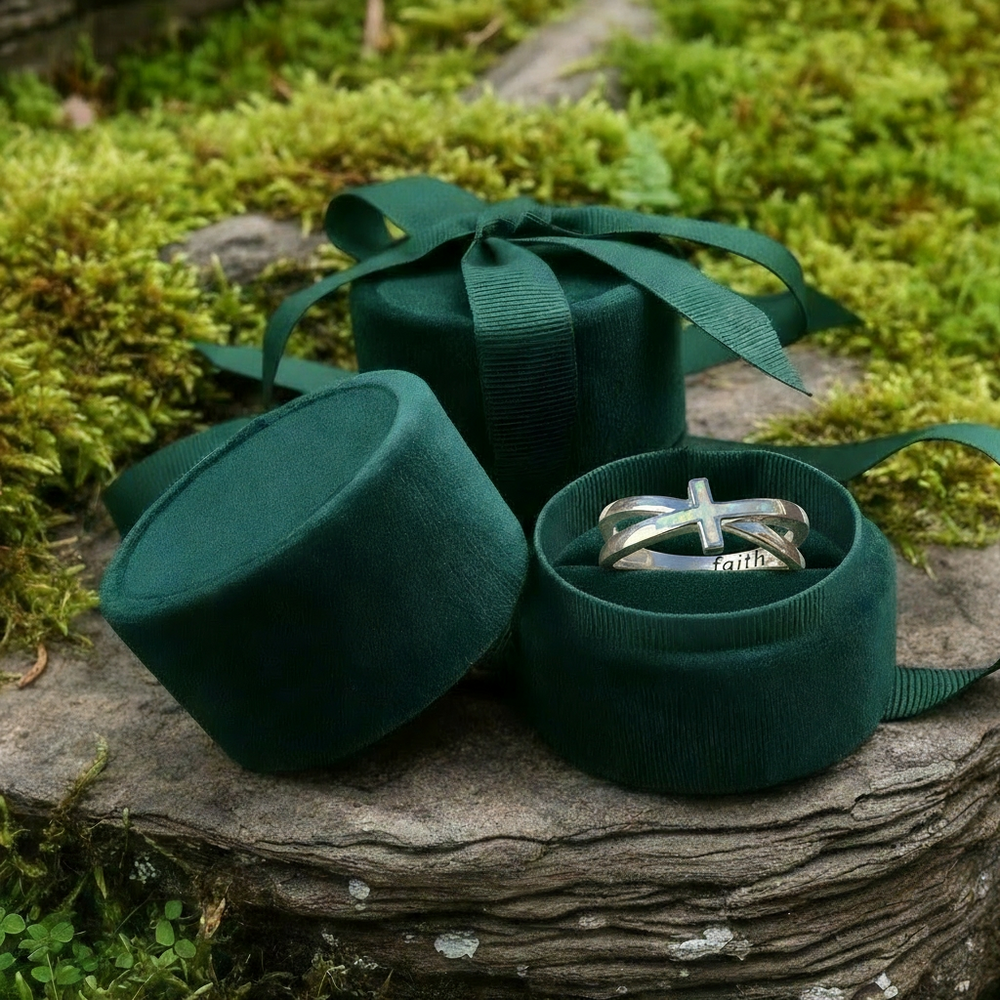 The opal cross ring nestled in an open round green velvet jewelry box with a ribbon-tied box behind it