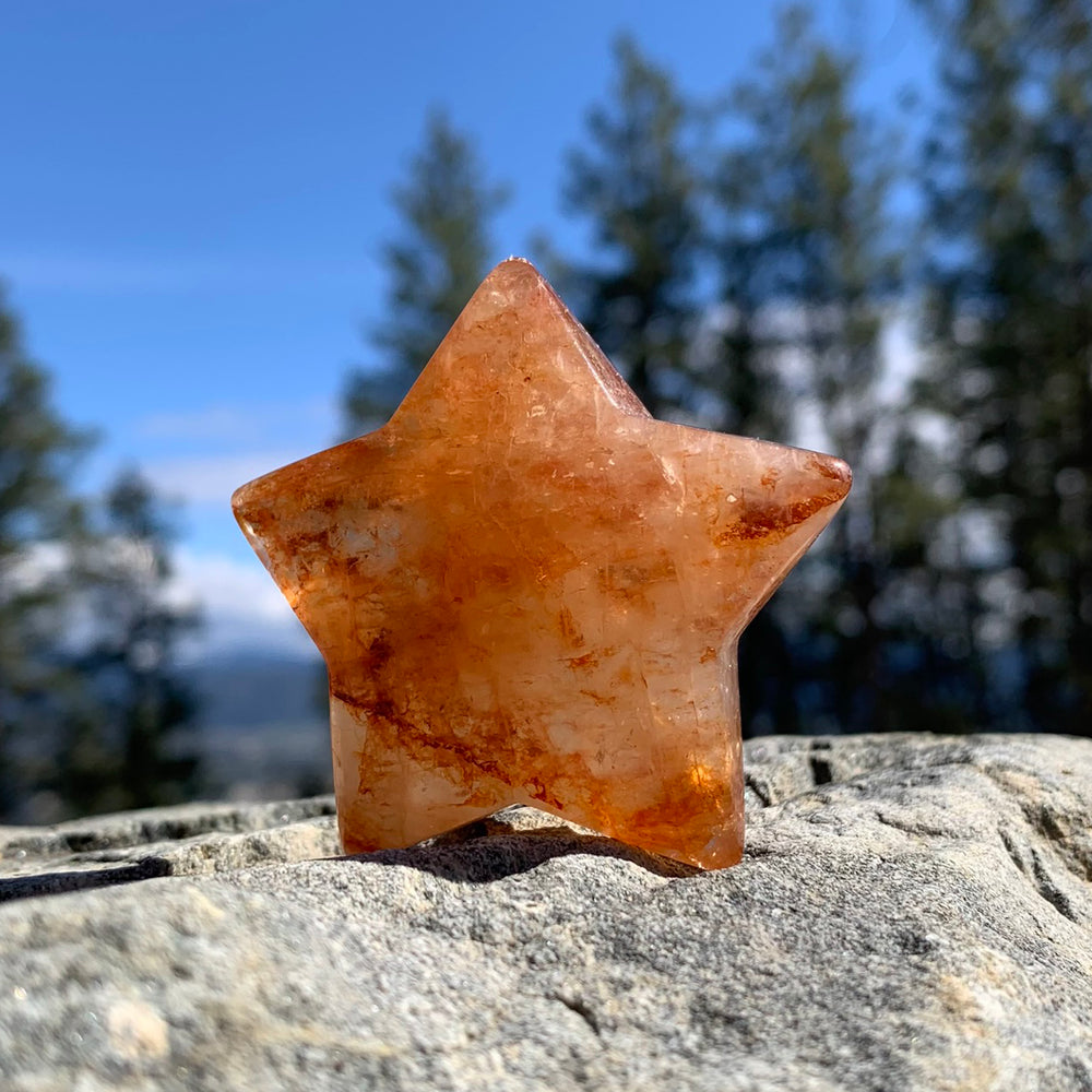 A polished five-pointed fire quartz star glowing orange with rust-red streaks, standing on a sunlit rock before pine trees