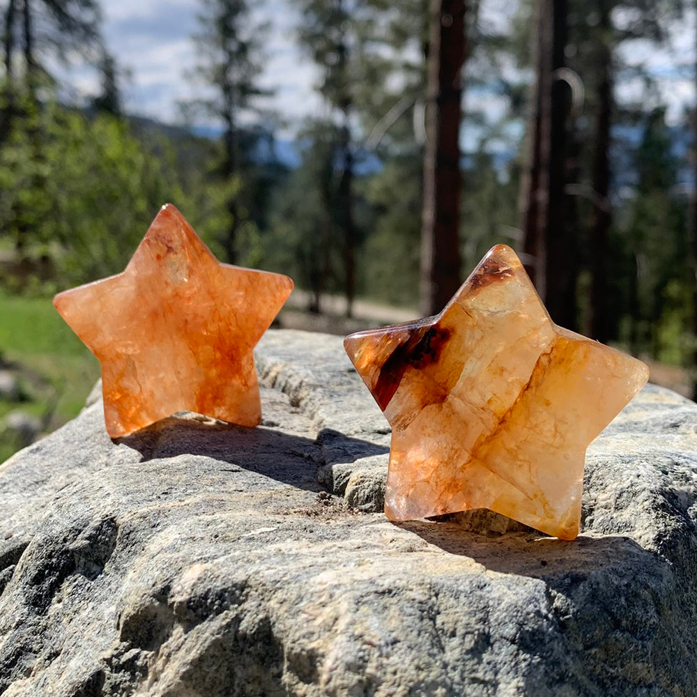 Two fire quartz stars on a rock among pine trees, one a soft even orange, one pale gold with a deep red streak