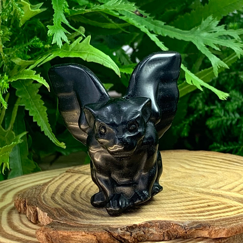 Black agate gargoyle with raised wings and a carved face, seated on a wooden slice in front of green ferns