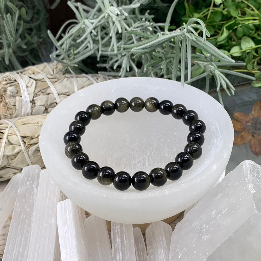 A single golden obsidian mala bracelet of round glossy black beads with a golden sheen, resting in a selenite bowl