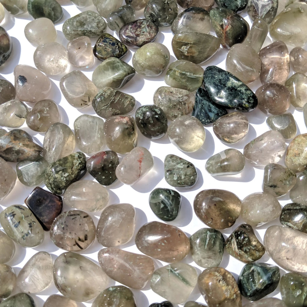 A scatter of tumbled green hair quartz stones, from clear and smoky to deep green, each with its own green threads and patches
