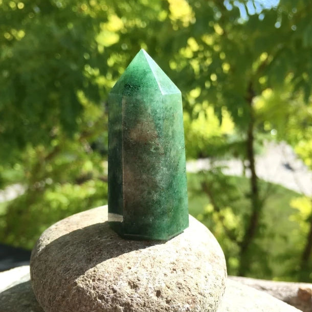 A polished green strawberry quartz point standing on a river stone in sunlight