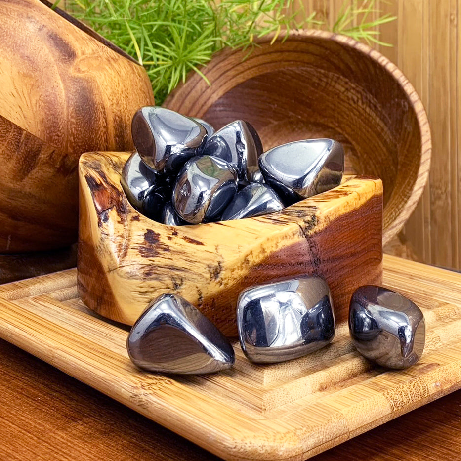 Glossy metallic hematite tumbled stones in a wooden bowl, with three more resting on a bamboo board in front