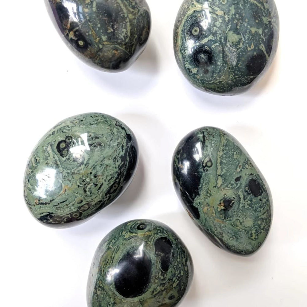 Five kambaba jasper palmstones on a white surface, each with its own mix of green, black and ringed orbs