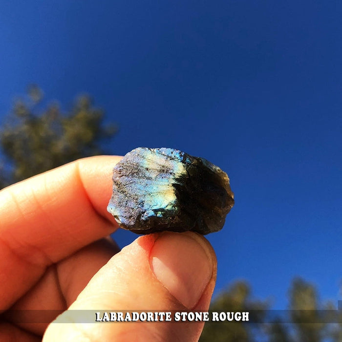 A rough piece of labradorite held between finger and thumb against a blue sky, showing a blue and gold flash