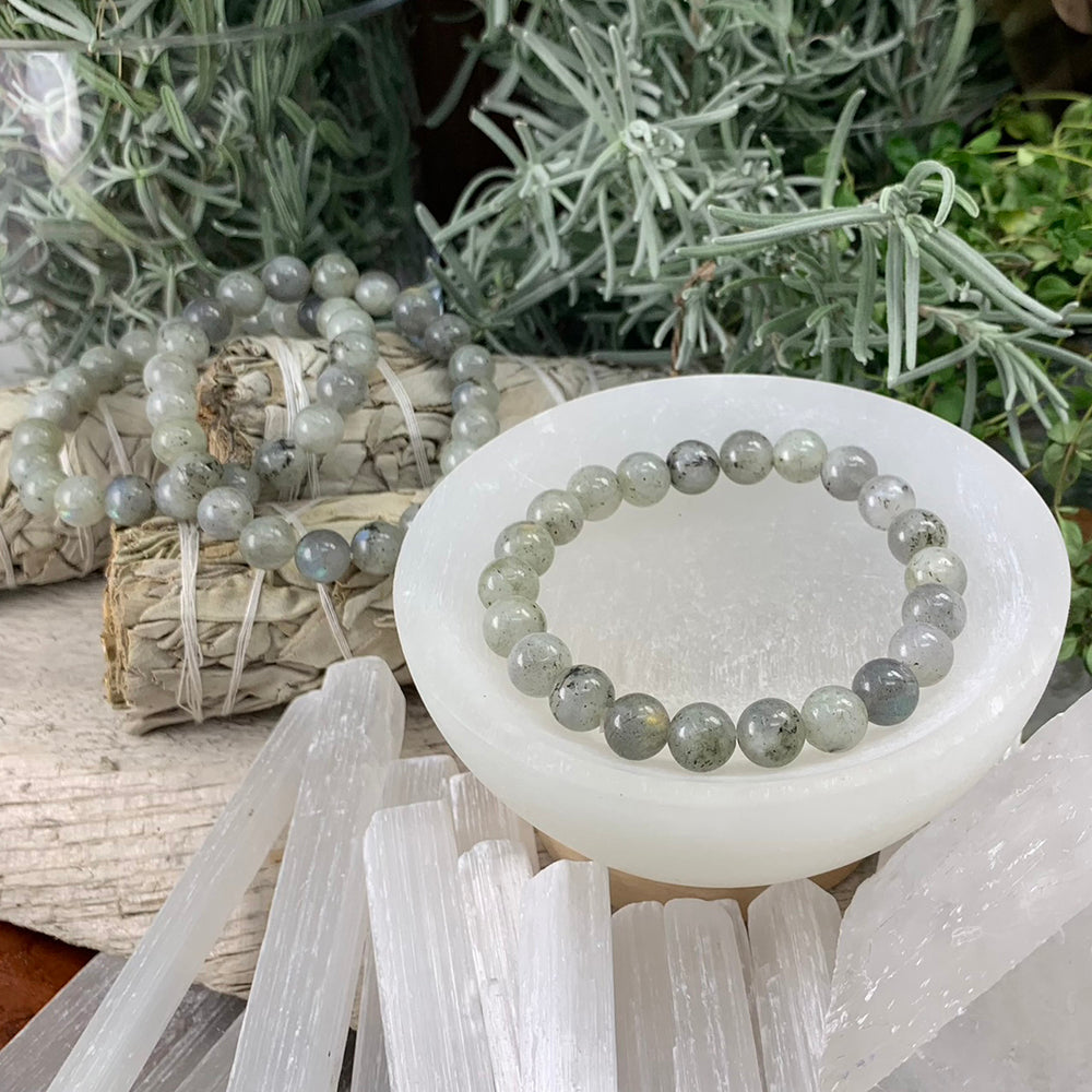 A labradorite mala bracelet of round grey-green beads with dark flecks resting in a white selenite bowl, with more bracelets draped behind