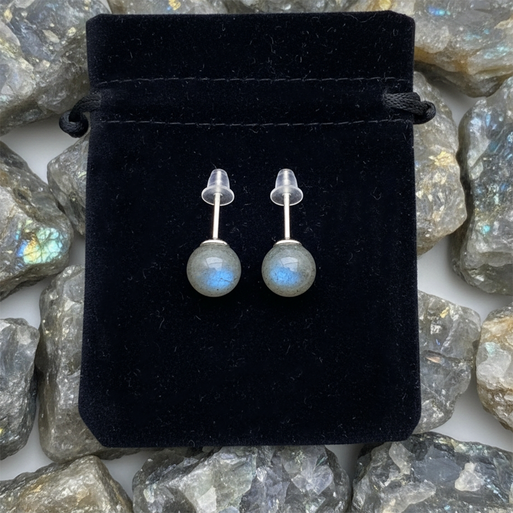 The pair of labradorite studs with their posts and clear backs, laid on a black velvet drawstring pouch among raw labradorite
