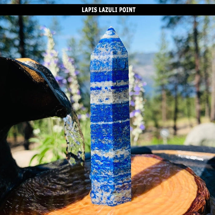 A tall faceted lapis lazuli point banded in blue and pale stone, standing outdoors by a small water feature