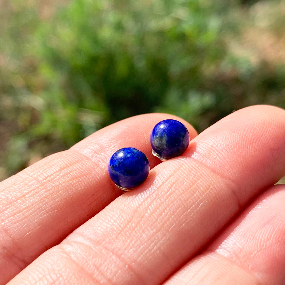 Two round polished lapis lazuli studs, deep blue with gold and white flecks, resting on fingertips in sunlight