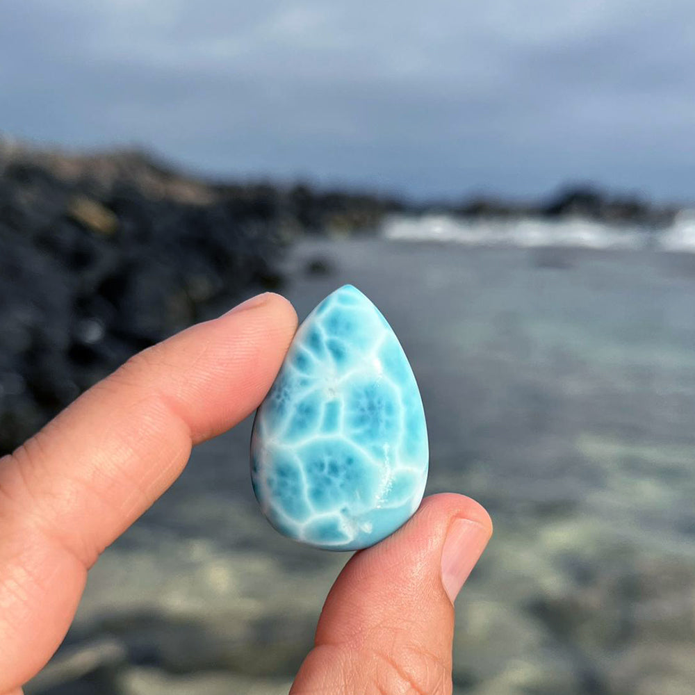 A Larimar teardrop held between two fingers in front of a Caribbean shoreline
