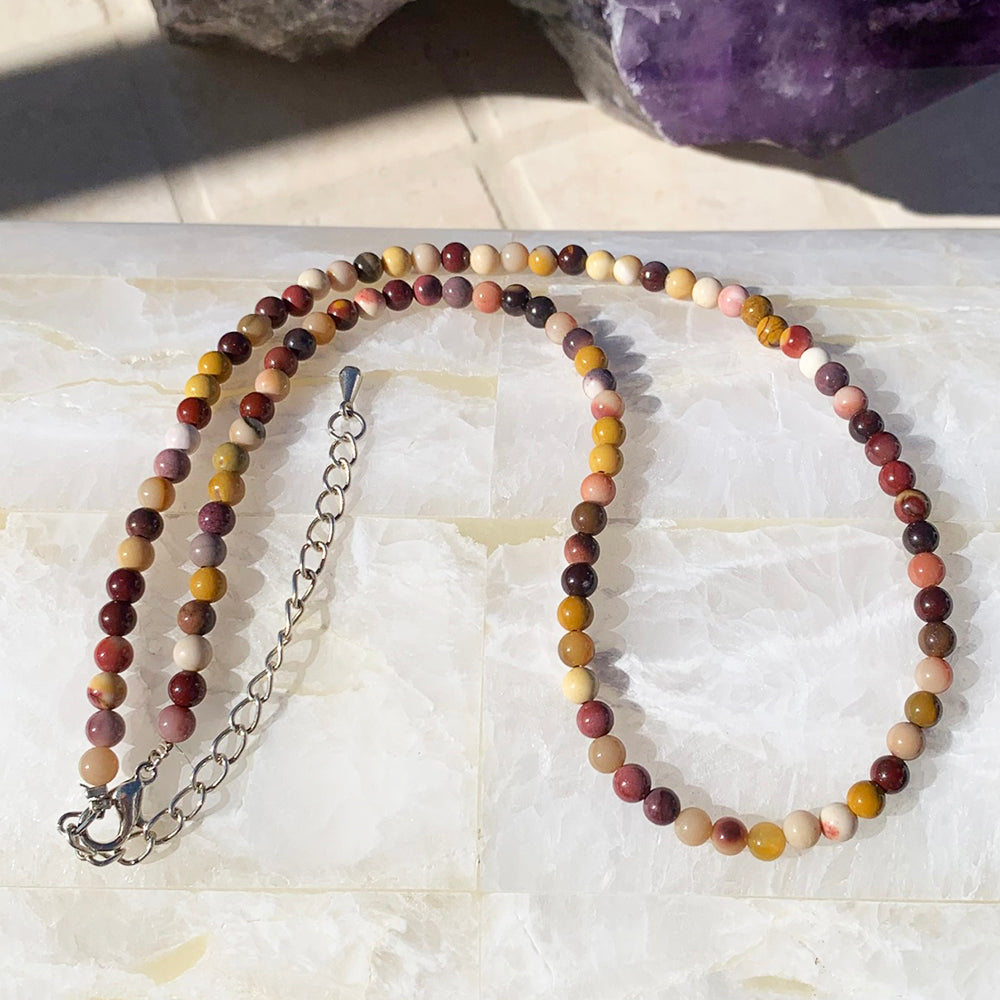 A mookaite bead necklace in crimson, ochre, gold and ivory, laid out with its silver clasp and extender chain on white stone