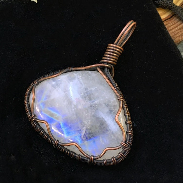 A copper wire-wrapped moonstone pendant on black velvet, flashing bright blue across the milky white stone