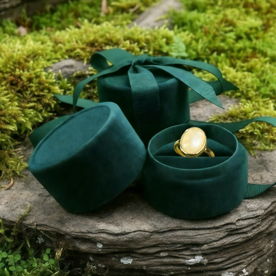 The moonstone and gold ring nestled in an open round green velvet ring box beside its lid and ribbon on a mossy stone