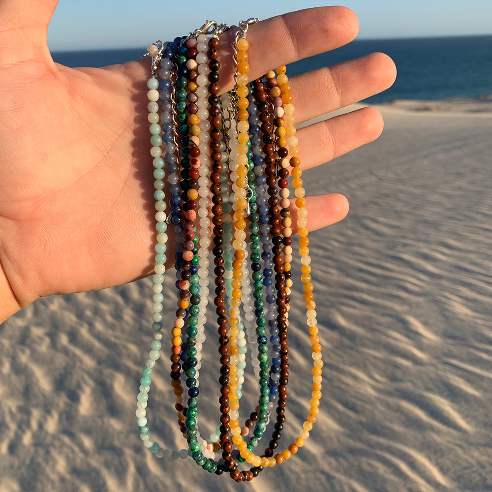 An open hand holding seven colourful chakra bead necklaces against a sand dune and the sea