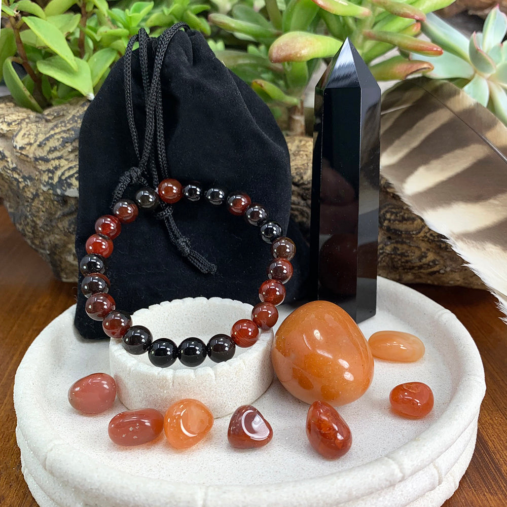 The full set on a white dish: a black obsidian point, a red aventurine ellipse stone, seven carnelian mini stones and a black and red agate bead bracelet draped over a black velvet pouch