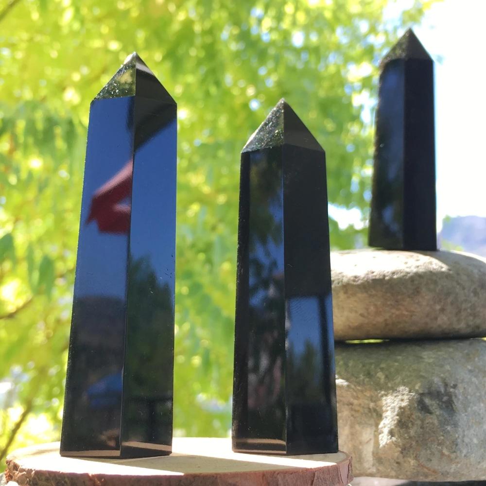 Three polished black obsidian points of different heights reflecting sunlight and leaves on their glossy faces