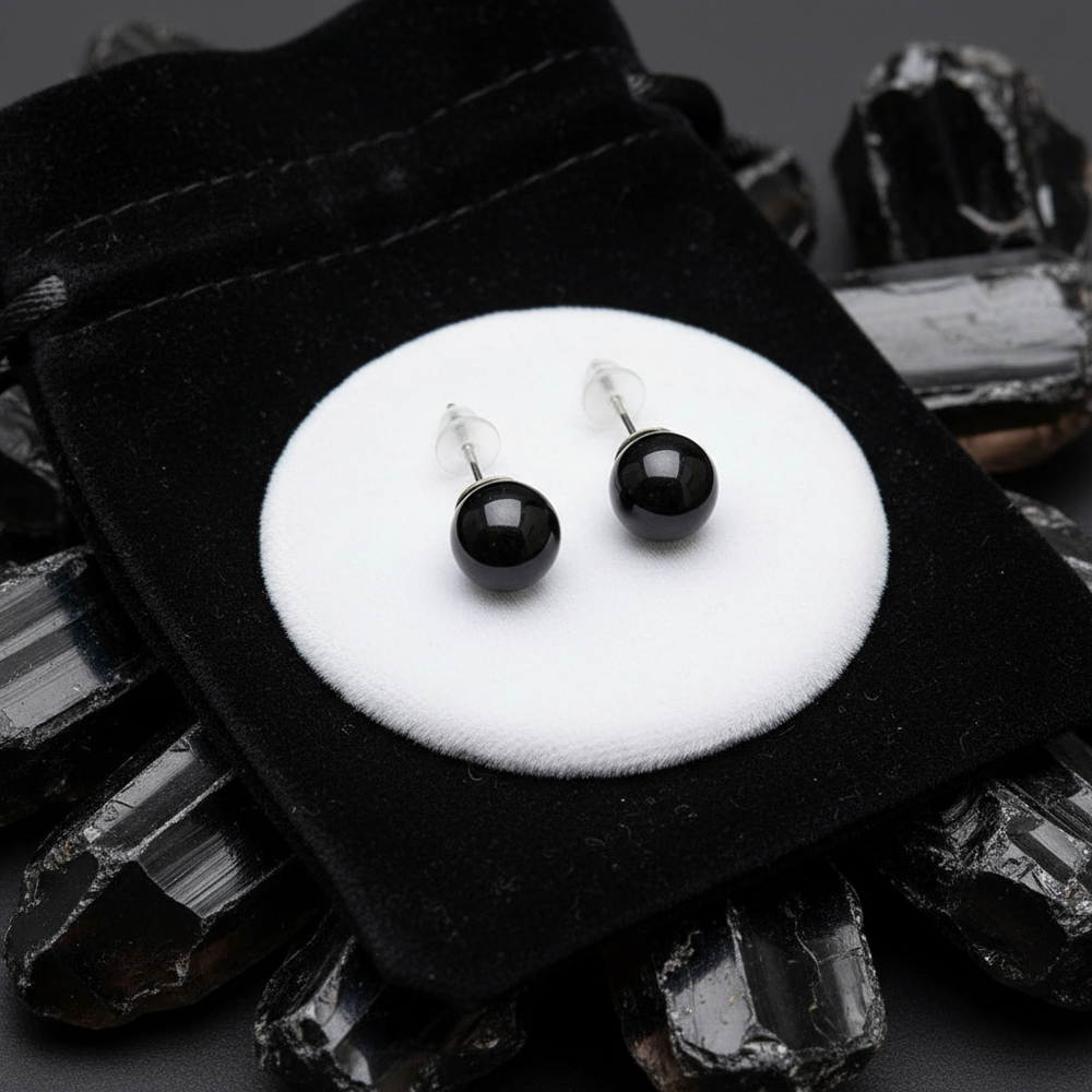 The pair of black obsidian studs with their posts and clear backs, set on a black velvet drawstring pouch among rough black stones