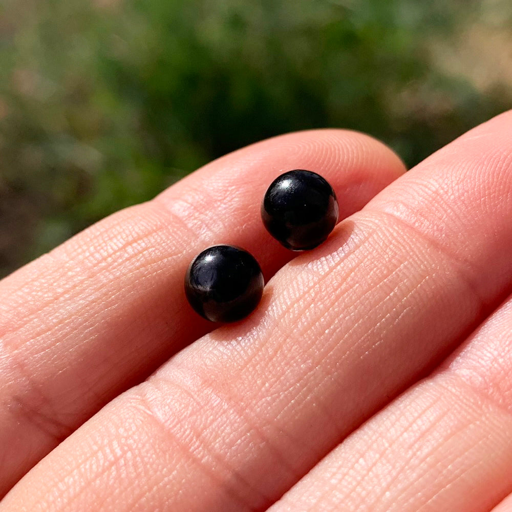 Two round, polished black obsidian studs resting on fingertips in sunlight