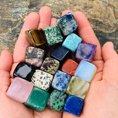 Two cupped hands holding nineteen colorful tumbled crystal cubes, including a milky opalite cube
