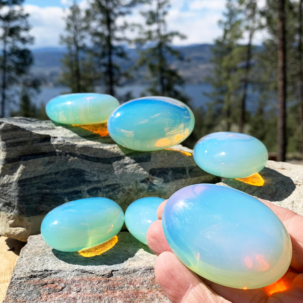 An opalite palmstone held in a hand outdoors, with more palmstones glowing on a rock behind it