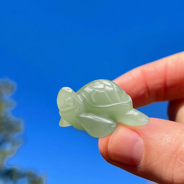 A small pale green hand-carved aventurine turtle held between fingertips against a blue sky