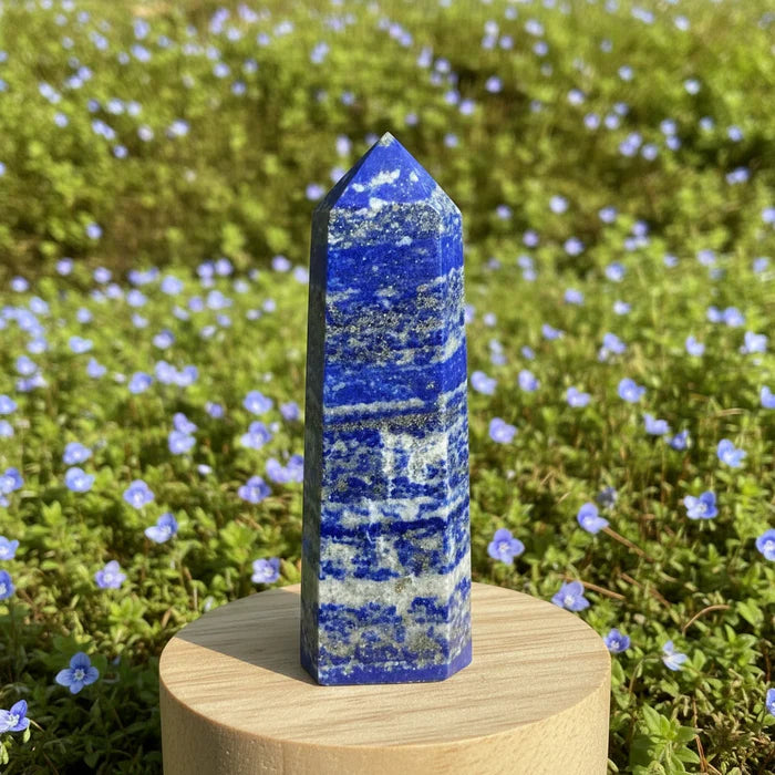 A tall lapis lazuli point with deep blue bands and golden flecks standing on a wooden round in a meadow of small blue flowers