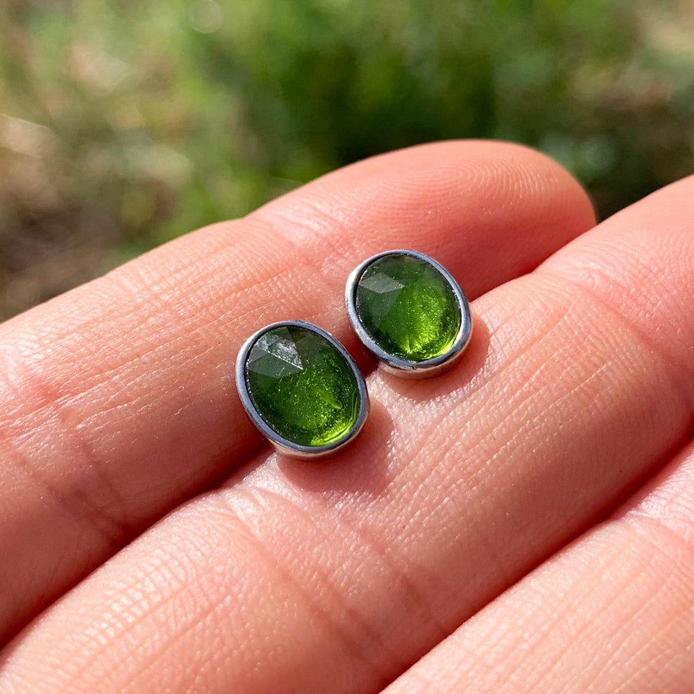 Two oval faceted peridot studs in sterling silver bezels resting on fingertips in sunlight